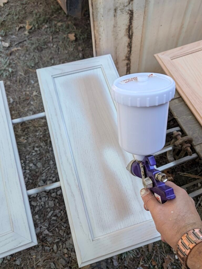 Using a purple gravity-feed HVLP spray gun to apply a light coat of white paint flat across an oak cabinet door